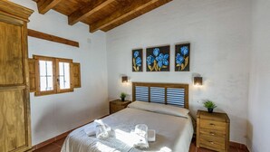 A bedroom with a wooden ceiling, a bed with a striped headboard, two bedside tables with lamps, and a wall with three framed floral paintings.