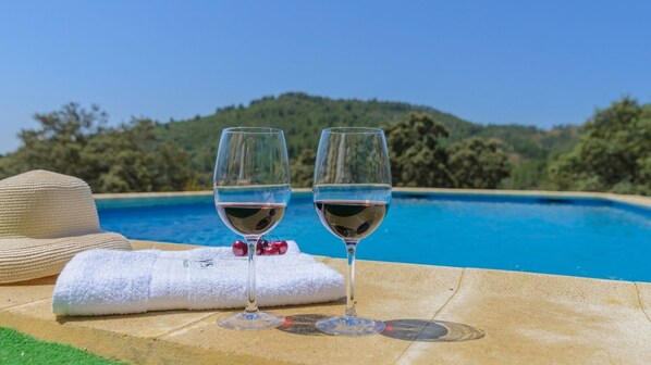 Dos copas de vino sobre una superficie de piedra junto a una piscina, con un sombrero y una toalla cerca.