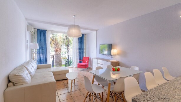 A modern living room with a glass dining table, white chairs, a red armchair, and a TV mounted on the wall. - Adosadas Lambda Deluxe Almuñécar by Ruralidays (Granada)