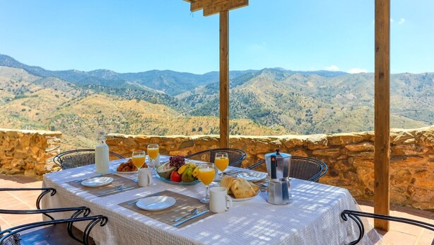 Una mesa dispuesta para el desayuno con vistas a la montaña, con platos, vasos y una cafetera.