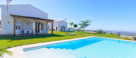 Blick vom Pool auf ein Haus, einen Pool, eine Terrasse mit Stühlen und den Ozean.