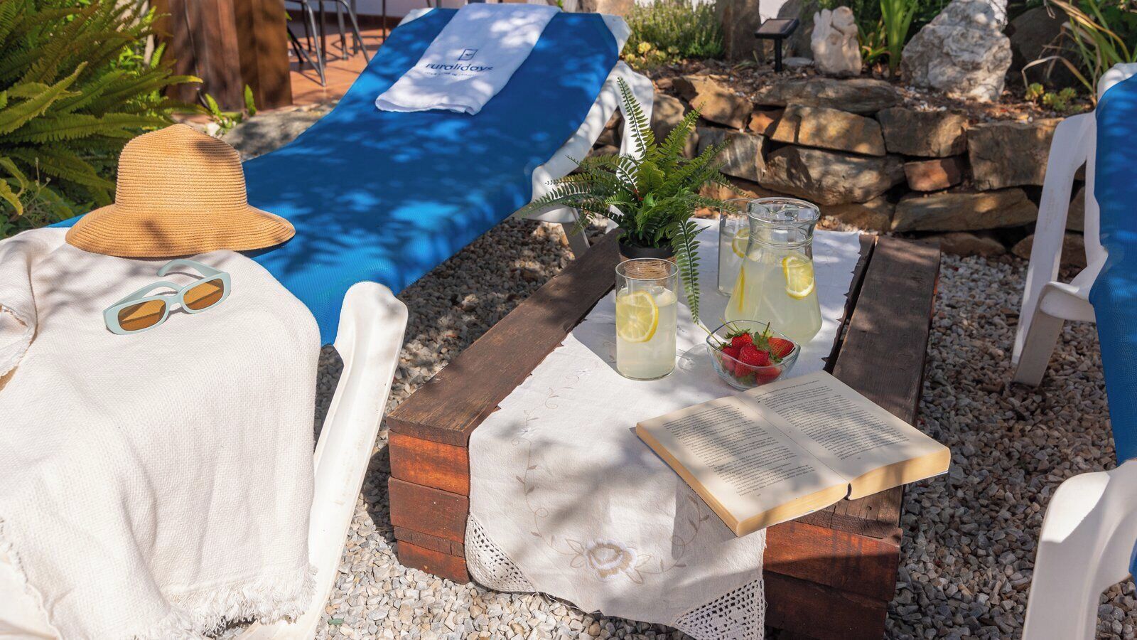 A sun lounger with a white towel, a straw hat, and sunglasses, a table with a book, a glass pitcher with lemonade, and a bowl of strawberries.