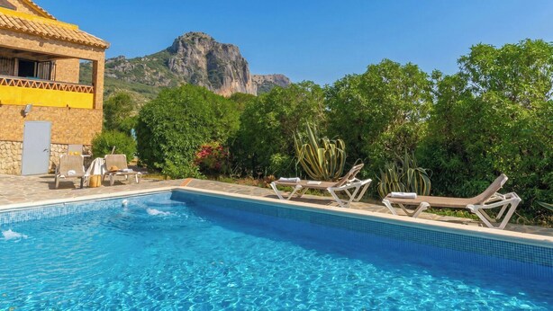 Una piscina con sillones de descanso, un edificio de piedra y una montaña al fondo.
