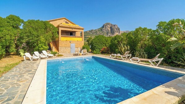 Una piscina con sillas de descanso, un edificio de piedra y una montaña al fondo.