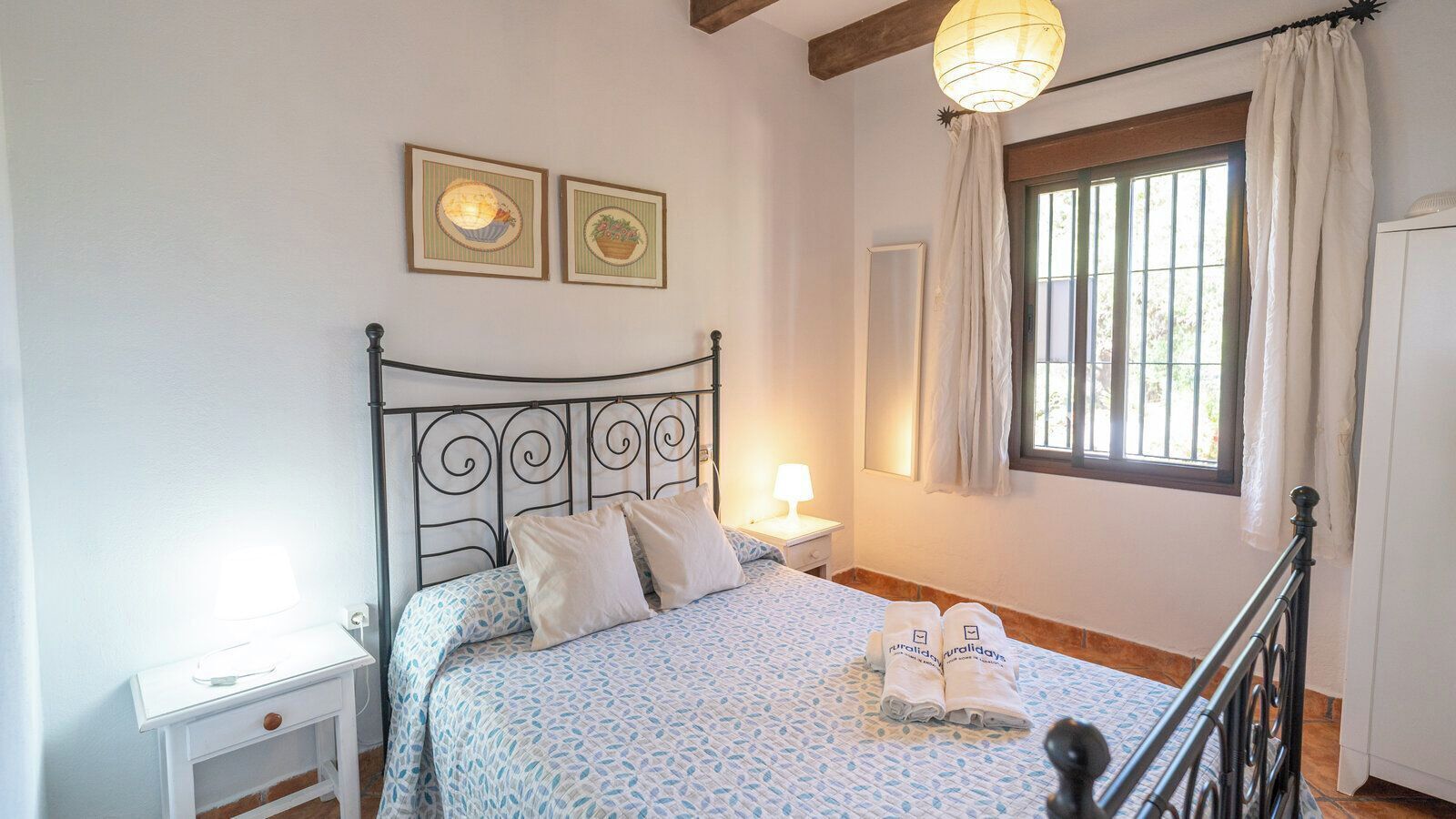 Une chambre à coucher comprenant un lit, une table de chevet, une lampe, une fenêtre avec des rideaux et deux tableaux accrochés au mur.