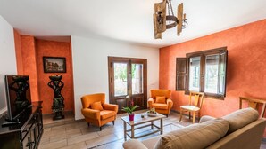 A living room with orange walls, two orange armchairs, a beige sofa, a wooden coffee table, and a TV stand.
