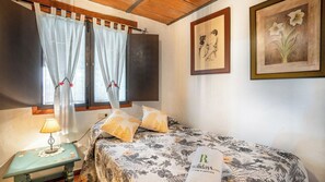 Un dormitorio con cama, una mesita de noche con lámpara, una ventana con cortinas y dos cuadros en la pared.