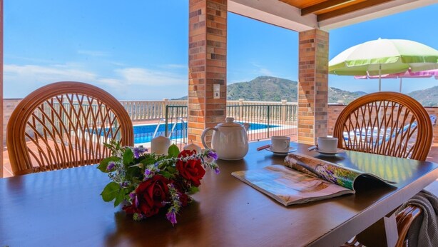 Une table avec un vase de fleurs, une théière et un magazine. Il y a deux chaises et un parapluie vert. On aperçoit une piscine au fond.