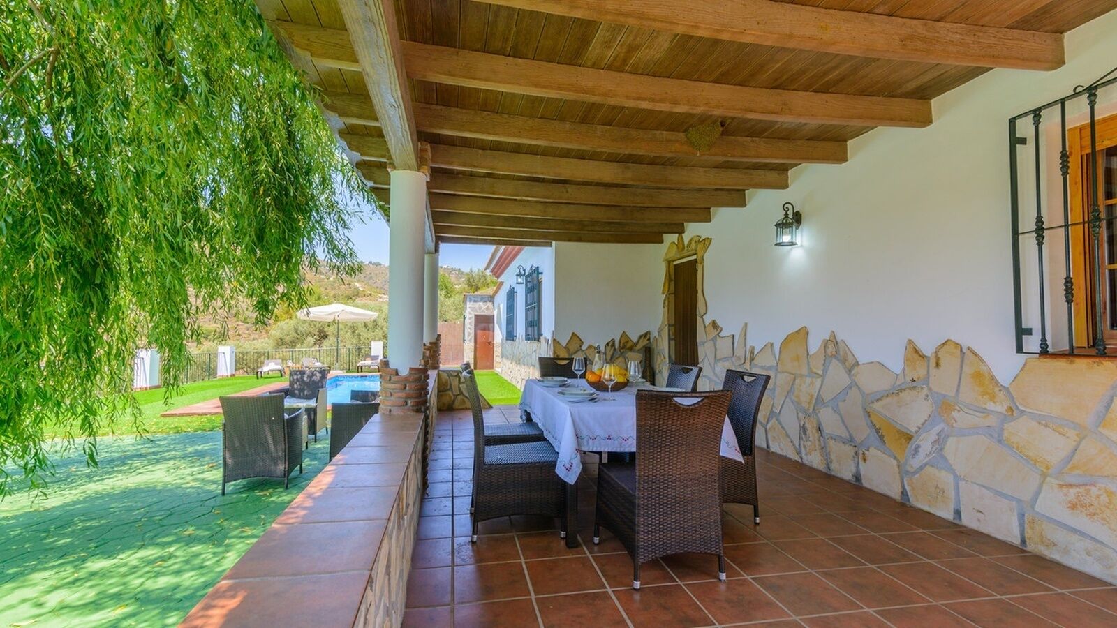 Un patio al aire libre con una mesa lista para comer, rodeado de muros de piedra y un techo de madera.