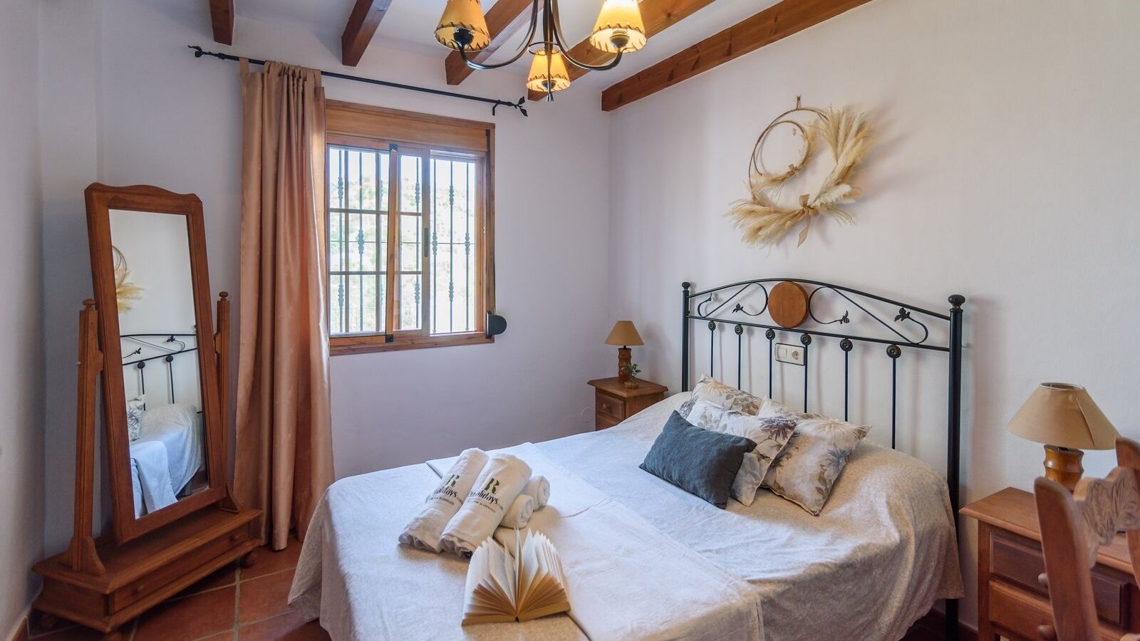 Une chambre à coucher comprenant un lit, une commode en bois, un miroir, une fenêtre avec des rideaux, une couronne décorative accrochée au mur et deux lampes de chevet.