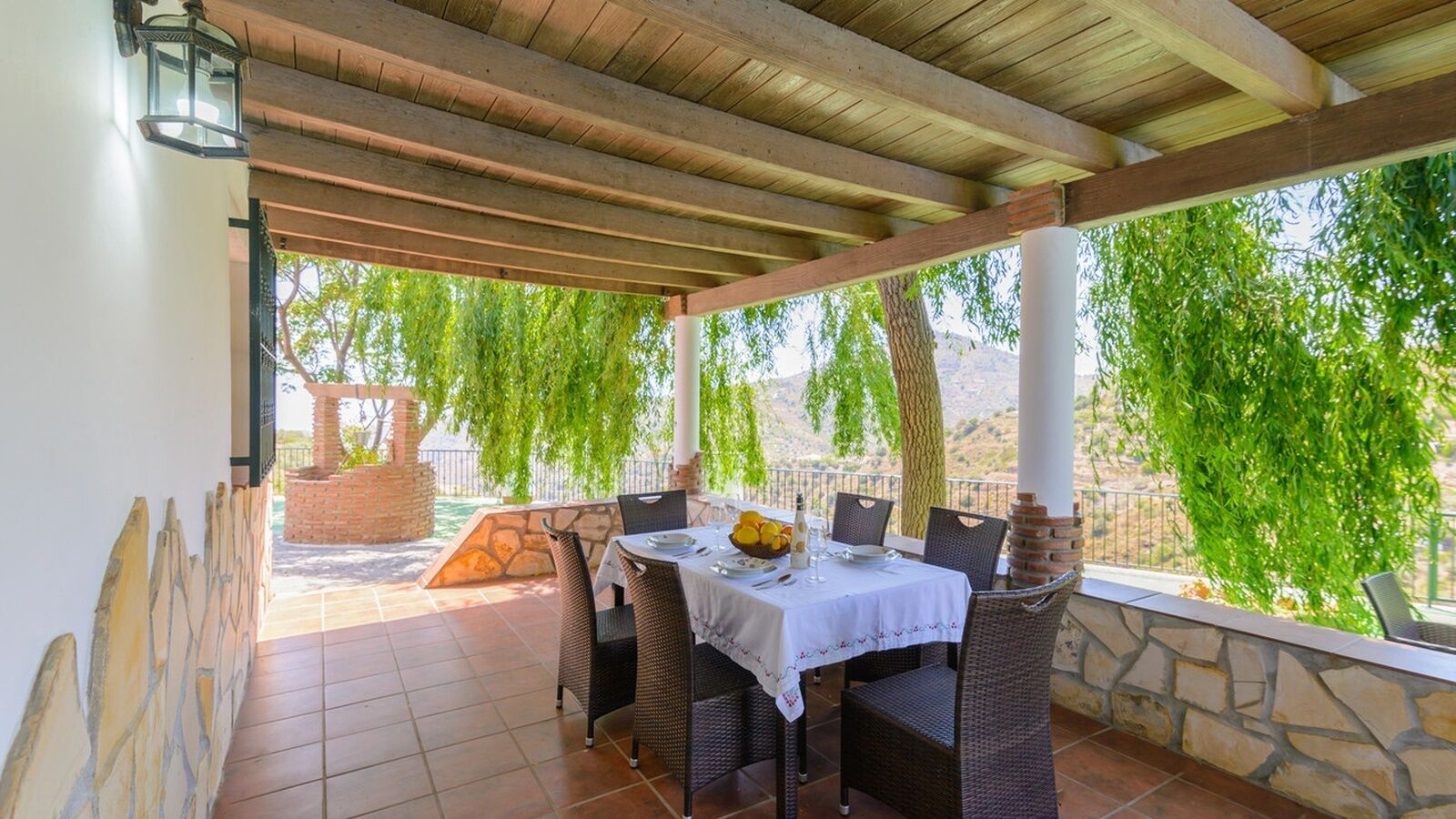 An outdoor patio with a table set for two, surrounded by wicker chairs, a stone wall, and a view of greenery and hills.