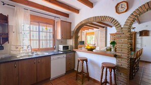 A kitchen with wooden cabinets, a white dishwasher, a microwave, and a brick archway leading to a dining area with a table and chairs. - Las Esparragueras Cómpeta by Ruralidays (Málaga)