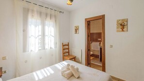 Une chambre à coucher comprenant un lit, une chaise, une porte en bois donnant sur une salle de bain et une fenêtre avec des rideaux légers.