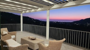 Un balcon doté de meubles en osier et offrant une vue sur un coucher de soleil au-dessus d’une ville.