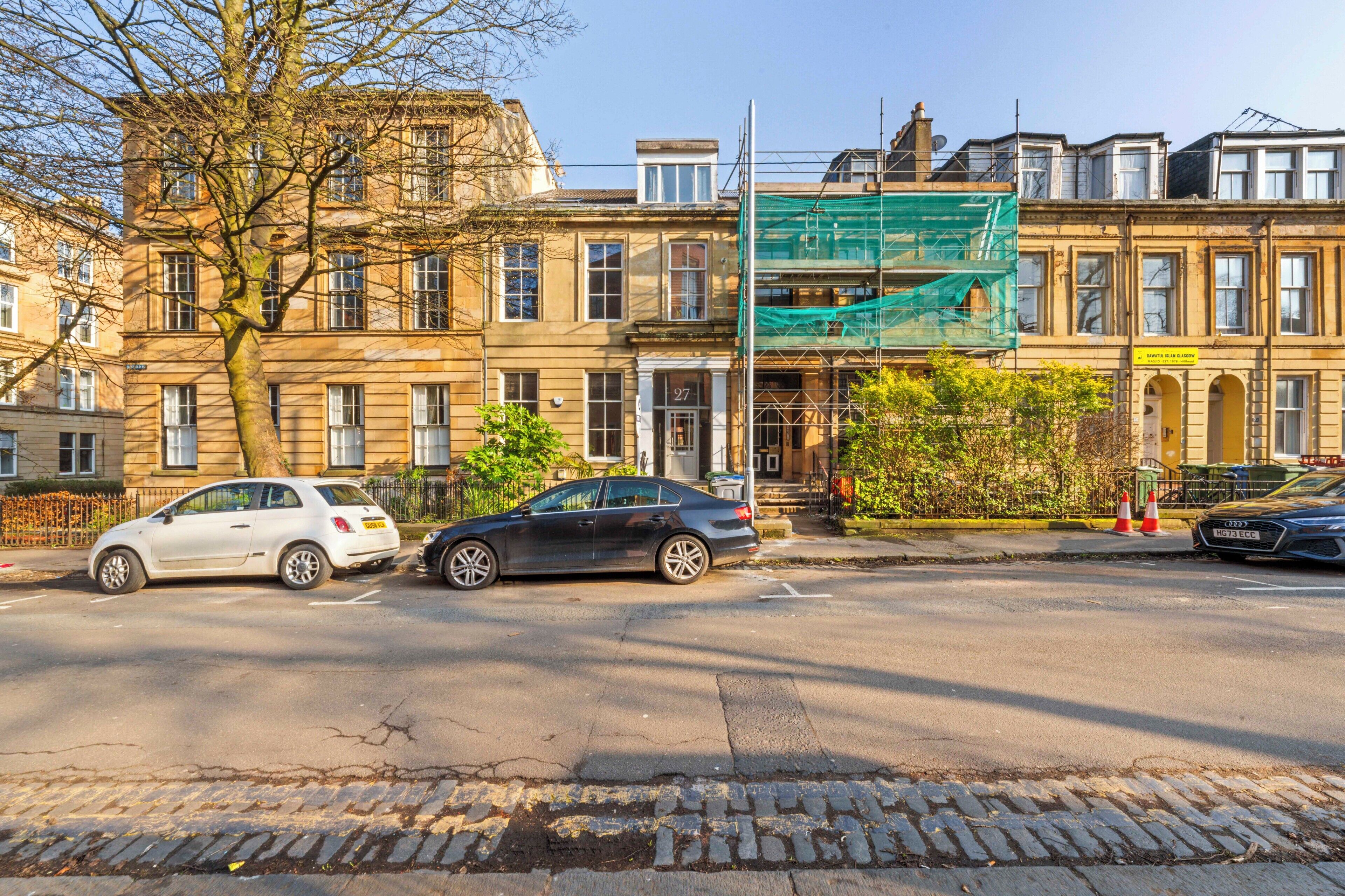 Une vue de rue avec deux voitures garées, un arbre et des bâtiments portant le numéro « 27 ».