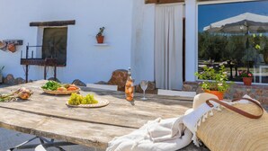 An outdoor dining area with a rustic wooden table, a bottle of orange juice, a basket of vegetables, and a woven bag.