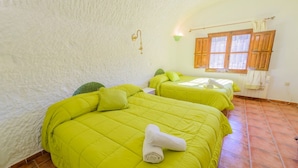 A room with two beds, each with bright green bedding and a white towel on the first bed. The room has a rustic, white textured wall and a wooden window with shutters. - Cuevas La Chumbera - Jazmines 9pax Fonelas by Ruralidays (Granada)