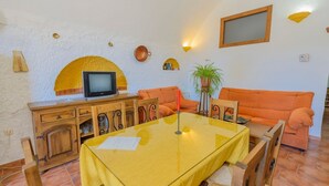 A dining area with a glass table, wooden chairs, an orange sofa, a television on a wooden cabinet, and decorative wall elements. - Cuevas La Chumbera - Jazmines 9pax Fonelas by Ruralidays (Granada)