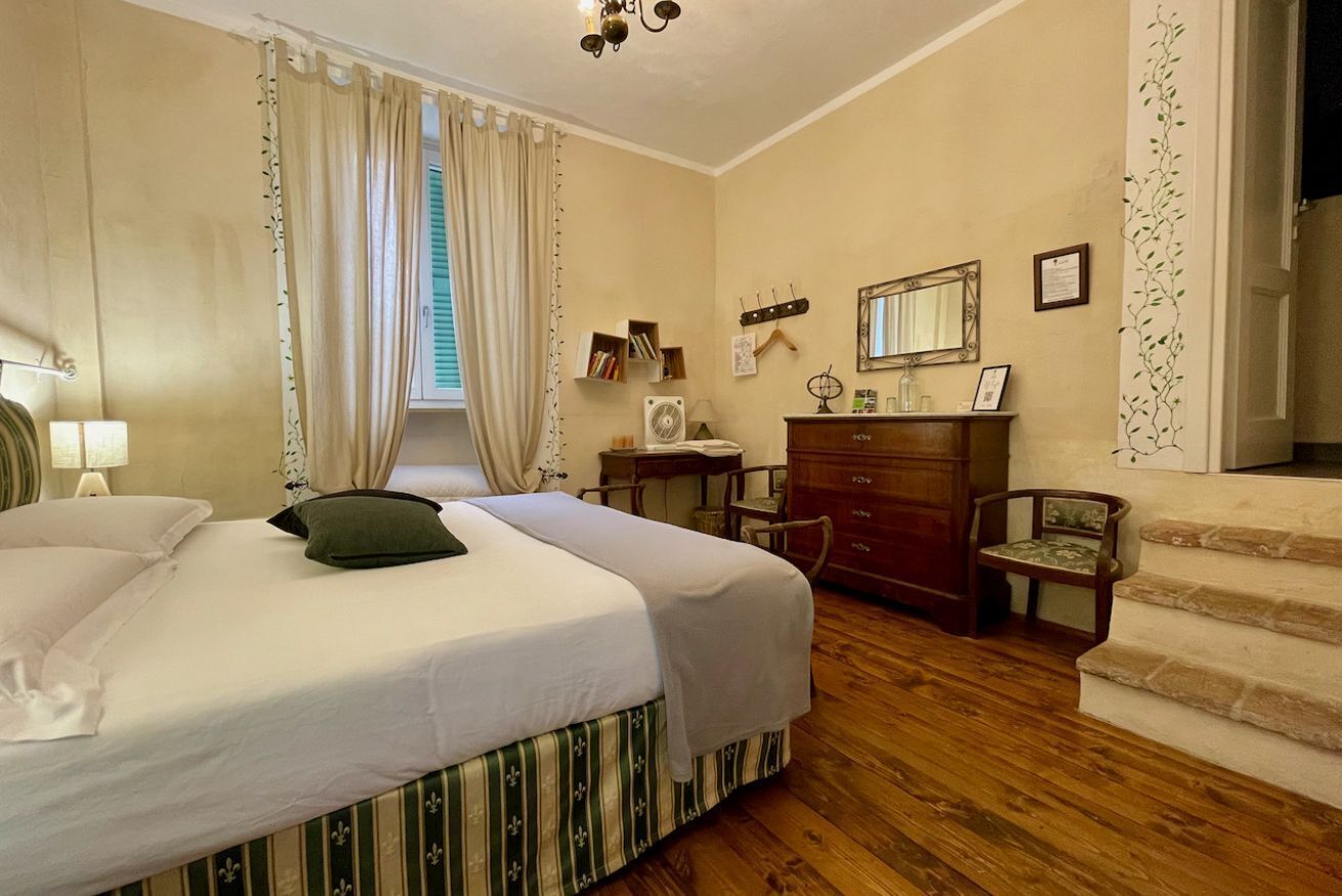 Quarto com cama, escrivaninha, cômoda, cadeira, espelho e janela com cortinas.