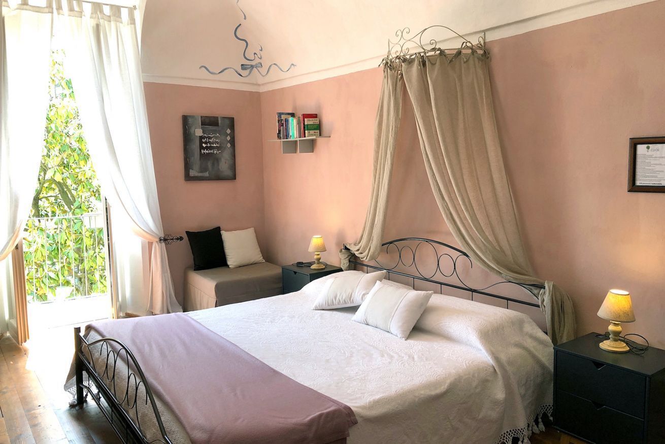 A bedroom with a bed, bedside tables, a nightstand, a window with curtains, and a wall-mounted shelf with books.