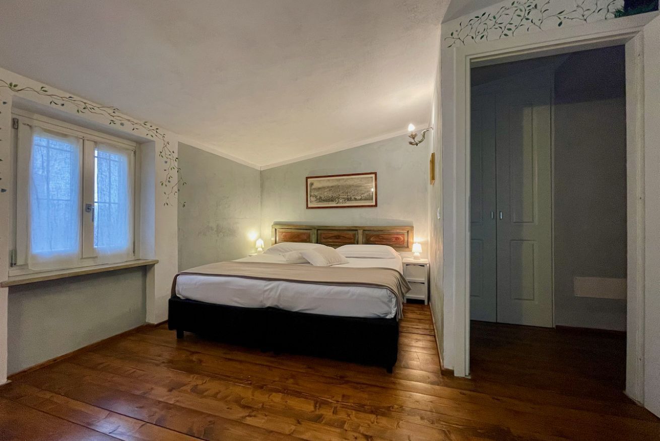 Una camera da letto con pavimento in legno, un letto con biancheria bianca, un comodino con una lampada, una finestra con tende e una porta che dà su un'altra stanza.
