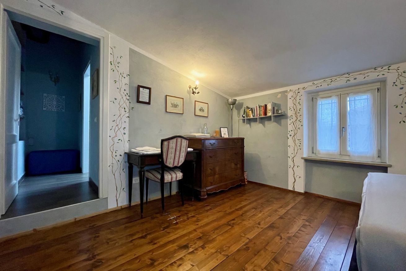 A room with wooden flooring, a desk with a chair, a dresser, a window with curtains, and a door leading to a blue room.