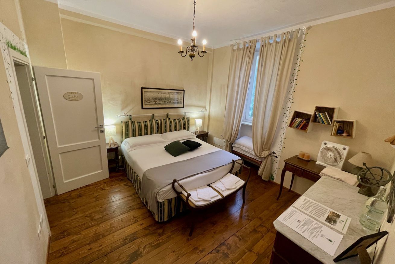 A room with a bed, a desk with a lamp, a chair, a window with curtains, a picture on the wall, and a door.