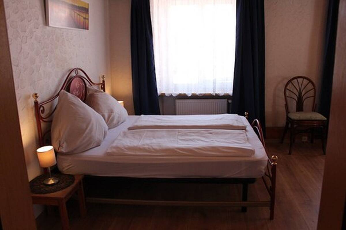 A single bed with a wooden frame, a bedside table with a lamp, a chair, and a window with curtains.