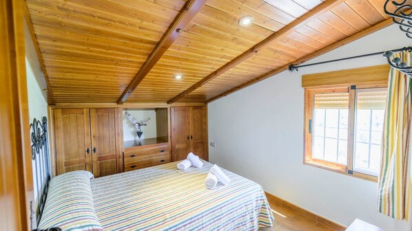 A bedroom with a wooden ceiling, a bed with striped bedding, a wooden wardrobe, and a window with blinds.