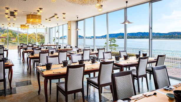 A restaurant with large windows offering a view of a lake and mountains. Tables are set with chairs and dining ware. - The Gran Resort Amanohashidate (Miyazu)