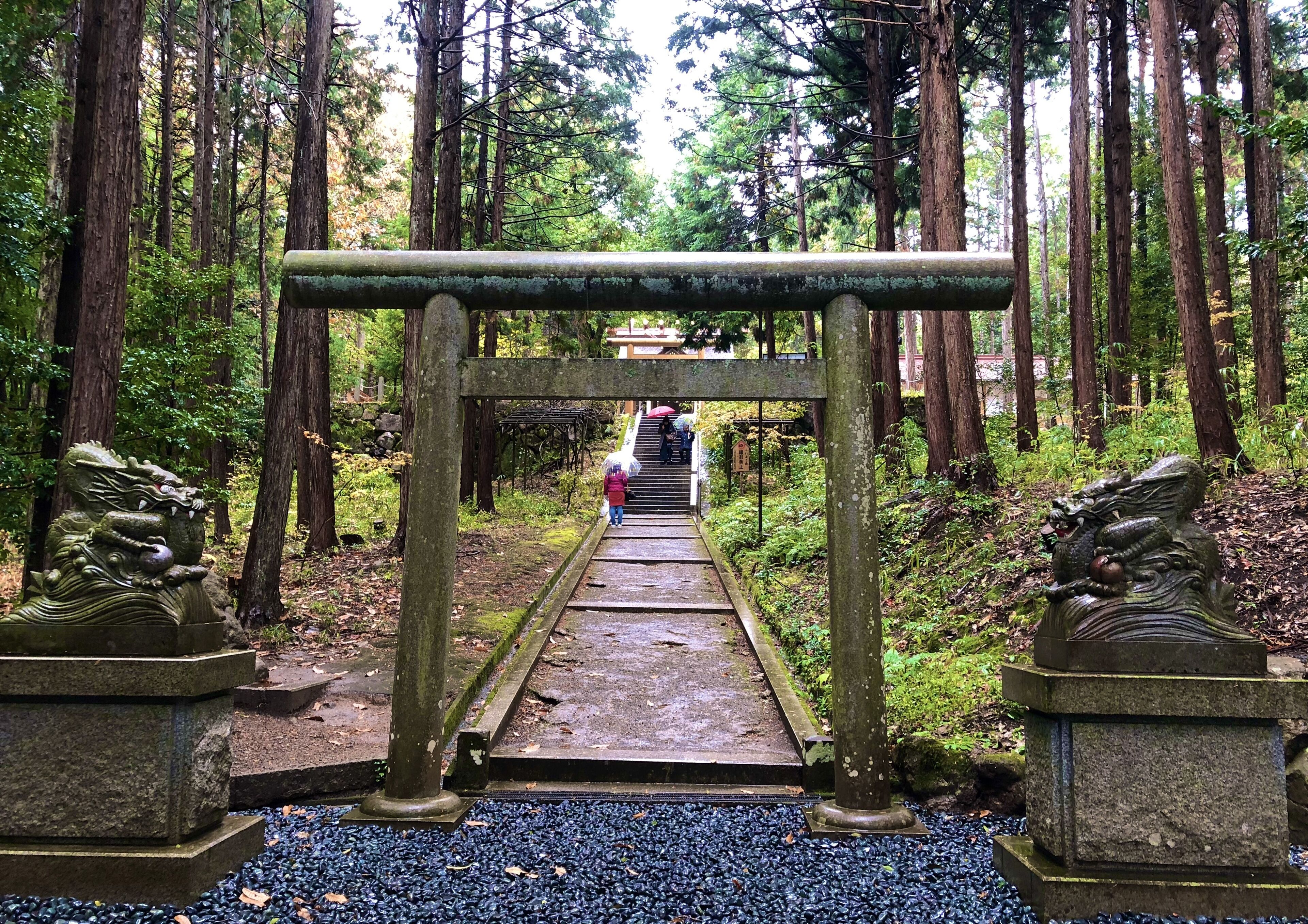 Un sendero de piedra serpentea por un bosque, flanqueado por estatuas de leones de piedra, y un pequeño santuario al final.