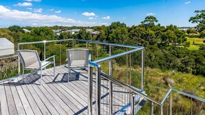 Eine Holzterrasse mit Metallgeländern und zwei Stühlen, mit Blick auf eine üppige grüne Landschaft mit Bäumen und Häusern in der Ferne.