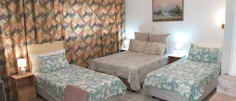 A hotel room with two single beds, a small table, a chair, a lamp, and a painting on the wall.
