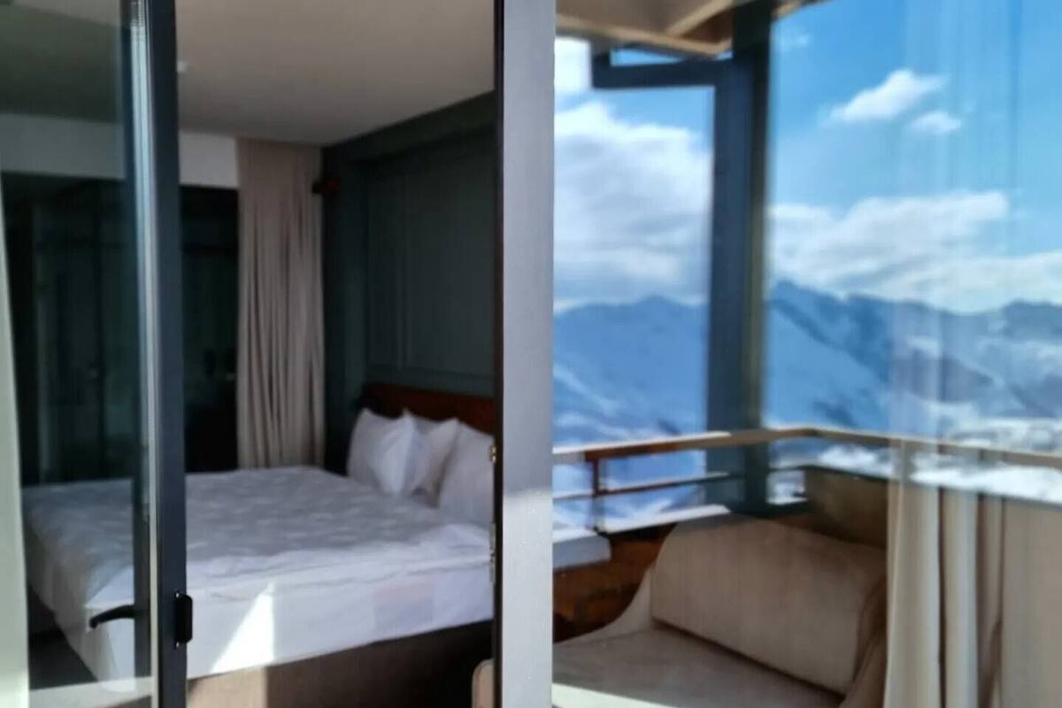 A modern hotel room with a large bed, a sofa, and a view of mountains through a large window.