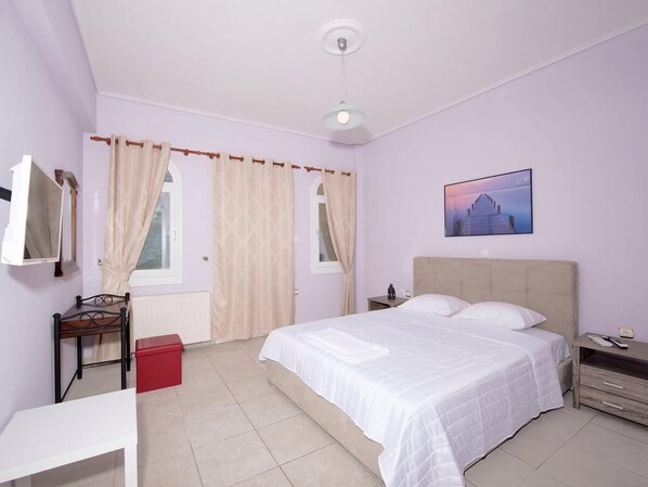 A bedroom with a bed, a bedside table, a nightstand, a window with curtains, a wall-mounted picture, and a radiator.