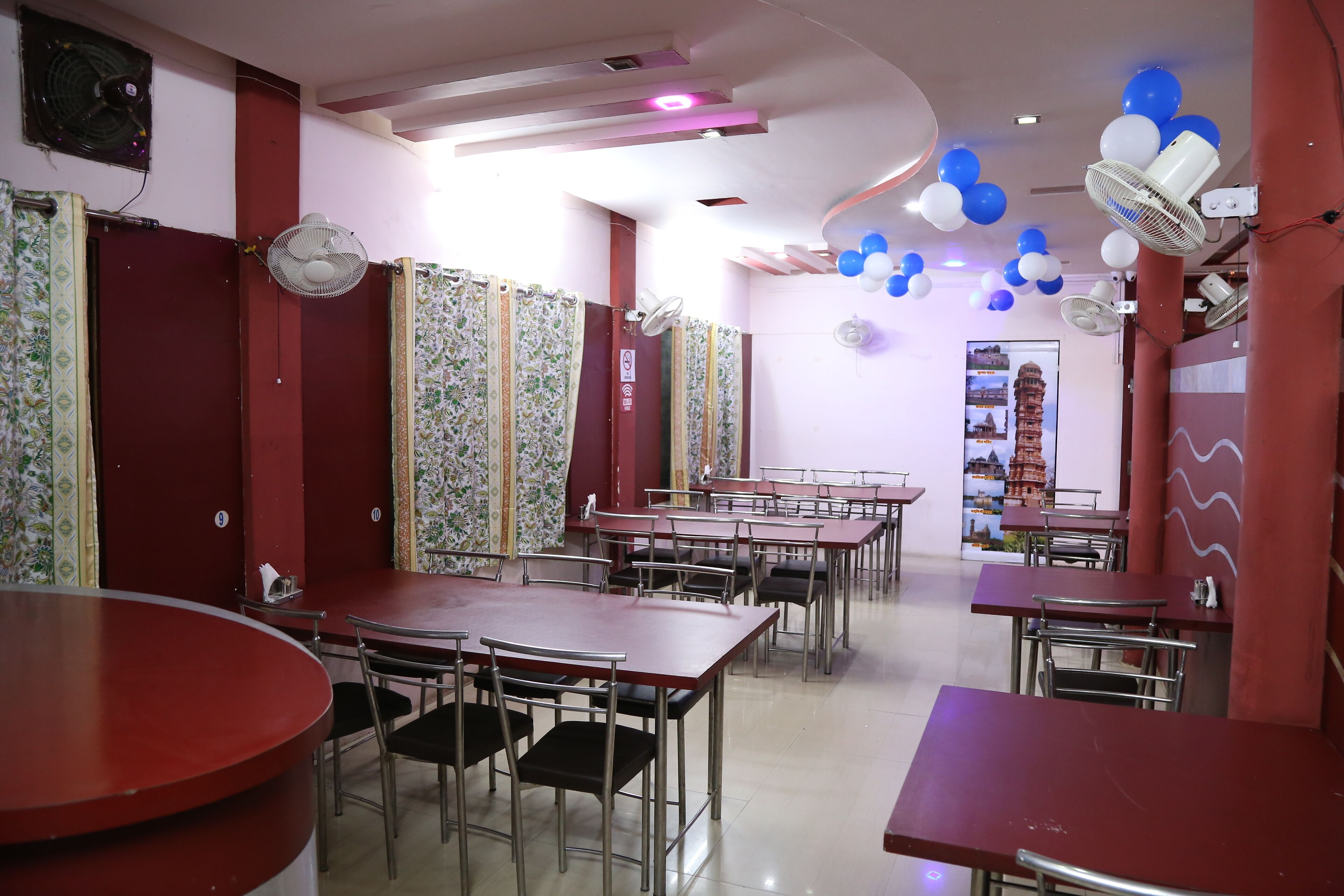 Un área de comedor con mesas y sillas rojas, ventiladores de techo y globos decorativos.