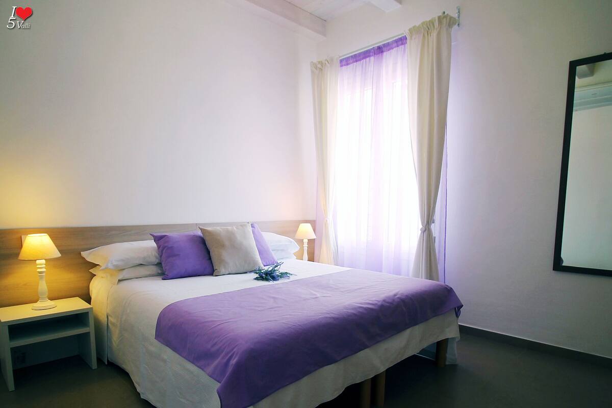 Una habitación de hotel con una cama, dos mesitas de noche con lámparas, una ventana con cortinas y un espejo en la pared.