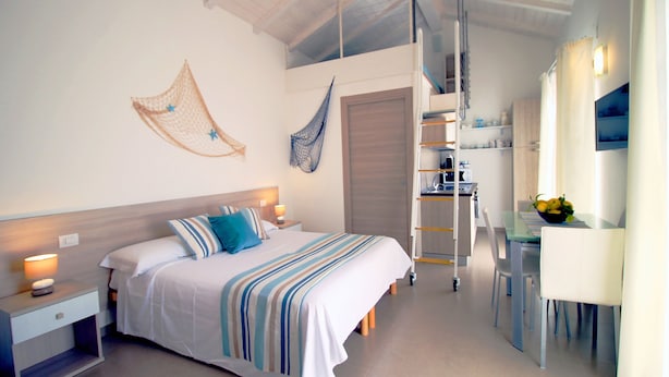 A bedroom with a bed, bedside tables, a desk, and a chair. There is a ladder leading to a lofted area with a kitchen.