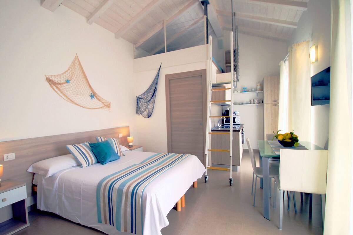 Un dormitorio con cama, mesitas de noche, un escritorio y una silla. Hay una escalera que lleva a una zona superior con cocina.