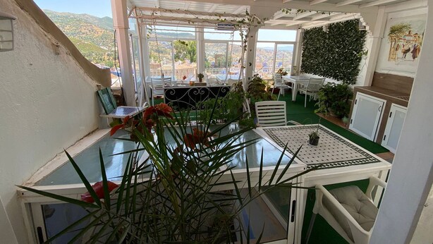 Un patio iluminado por el sol con una mesa y sillas, rodeado de plantas en macetas y vistas a colinas distantes.