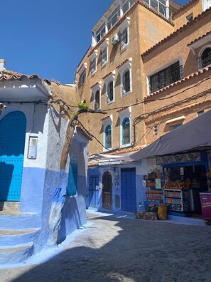 A narrow street with buildings painted in blue and white, featuring arched doorways and windows, and a small shop with various goods displayed outside. - Casa Elias (Chefchaouen)