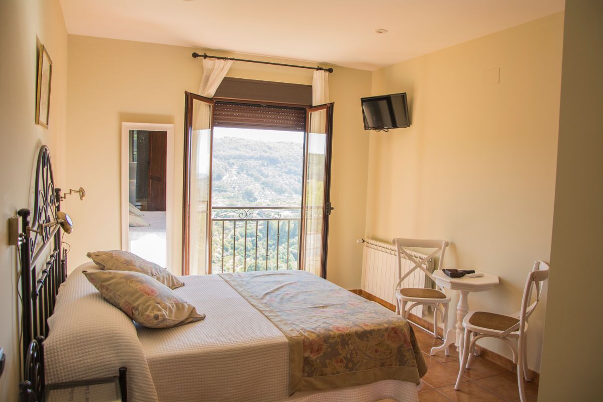 Un dormitorio con cama, una silla, una mesita, televisor y balcón con vistas.