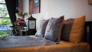 A neatly made bed with a dark bedspread and pillows, a small table with a lamp and a vase of flowers, and a window with a view of greenery. - Oimho Beach House Apartment Beach Front (Koh Samui)