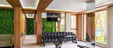 A modern gym with wooden beams, a wall-mounted TV, a bench press, dumbbells, and a green wall.