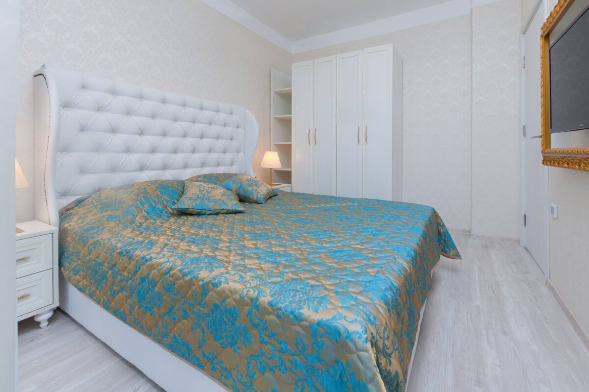 Un dormitorio con cabecera capitonada, cama con colcha estampada, mesita de noche con lámpara y armario con espejo.