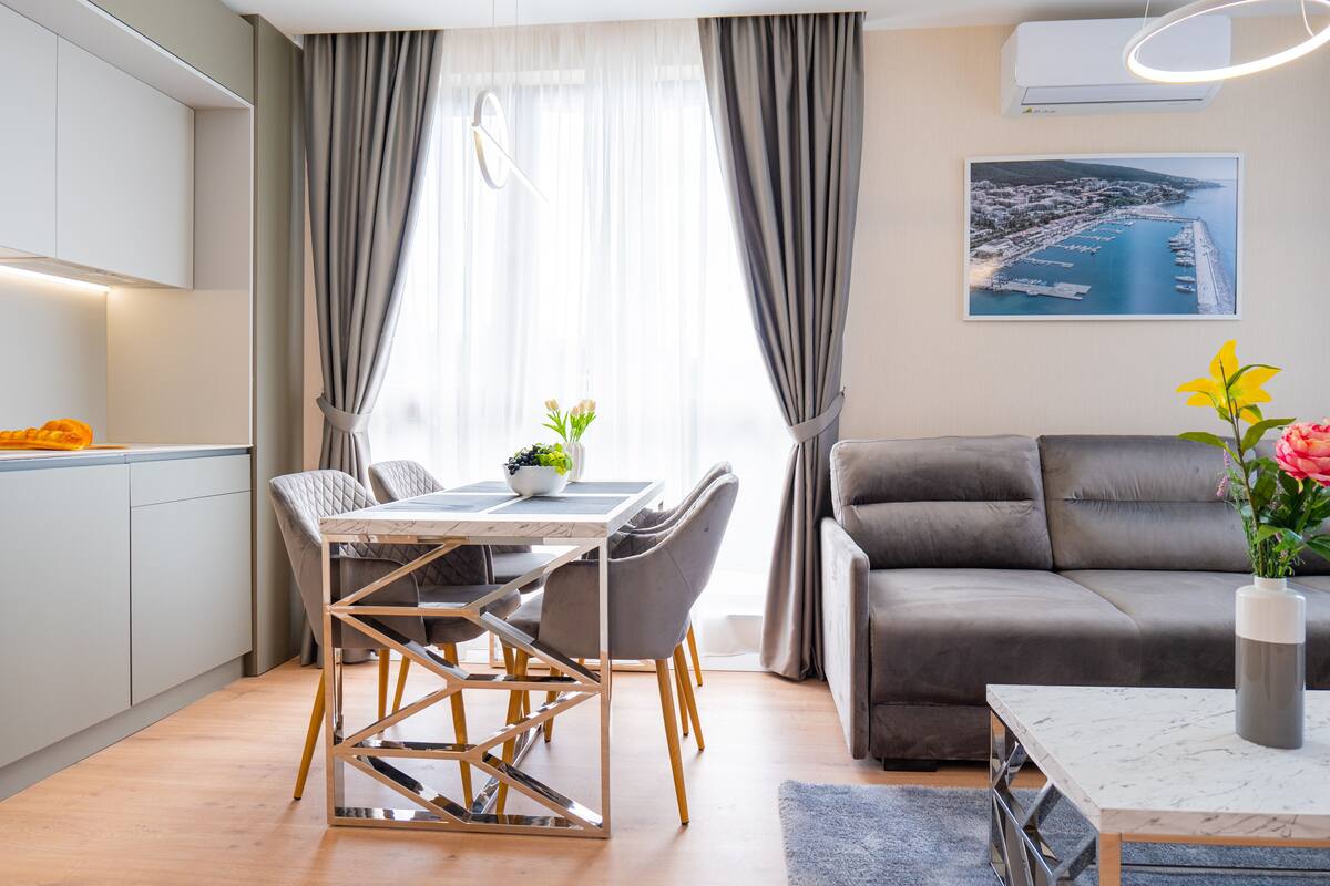 Una sala de estar moderna con un sofá, una mesa de comedor y sillas. Hay una ventana con cortinas, un cuadro en la pared y un florero con flores.