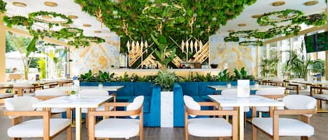 Un restaurante moderno con una zona de estar central, rodeado de mesas y sillas, y un techo decorado con follaje.