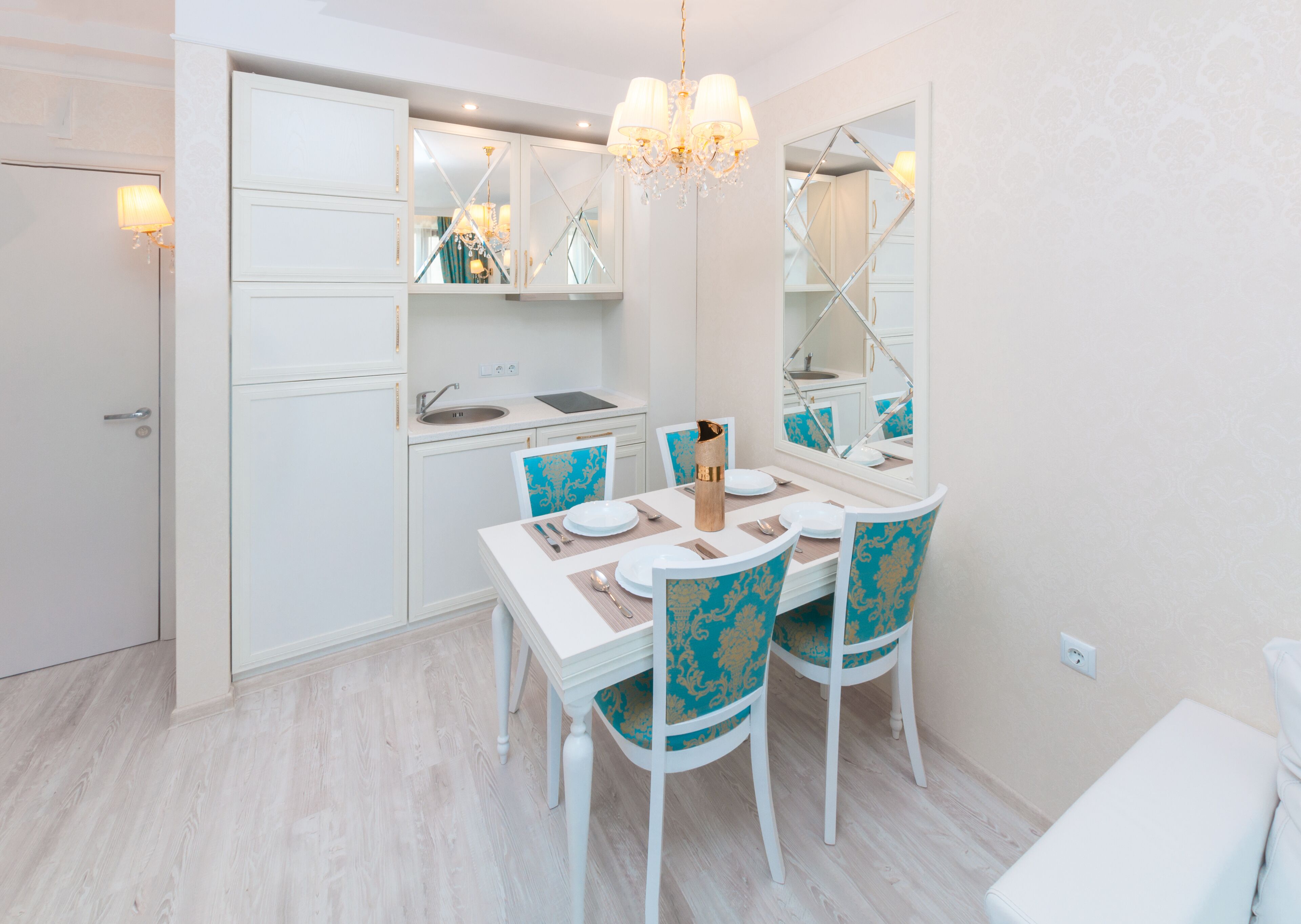 Un comedor moderno con una mesa blanca dispuesta para cuatro, sillas azules con detalles dorados, una araña y un espejo grande sobre la mesa.