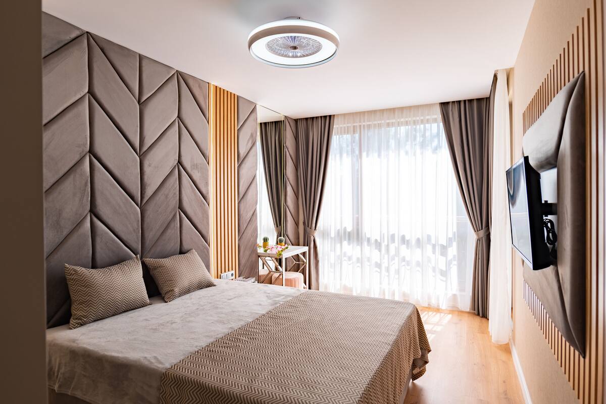 Un hotel moderno con una cama grande, un televisor montado en la pared y vistas de la ciudad a través de amplias ventanas.