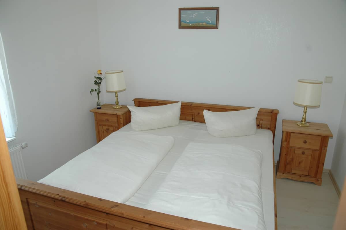 Una habitación con una cama individual, cabecera de madera, dos mesitas de noche con lámparas, un pequeño jarrón con flores y un cuadro en la pared.